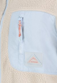 Fleecejacka med ett ljusblått tyg och beige strukturerad fleece. Har en dragkedjeficka med en gummiloggopatch och kontrasterande dragsko.