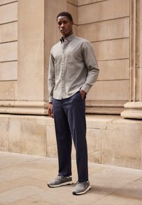 Grå button-up skjorte med en lomme, parret med mørkeblå stribede bukser og grå sneakers, stående foran en neutral baggrund.