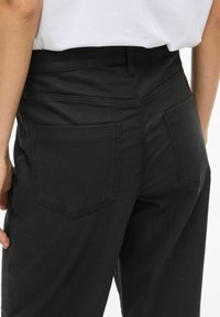 Juodi džinsiniai kelnės su prigludusia juosmens dalimi, dviem galinėmis kišenėmis ir matomomis siūlių detalėmis. Audinys turi lygų paviršių ir struktūruotą dizainą.