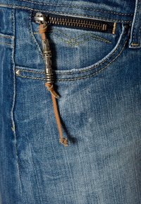 Gros plan sur la poche d'un jean en denim bleu avec une fermeture éclair métallique et une lanière en cuir marron décorative ornée de perles argentées suspendues à la tirette.