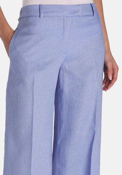 Vrouw draagt lichtblauwe, wijd uitlopende broek met hoge taille, zijzakken en een verborgen voorkantsluiting, kort geknipt ter hoogte van de taille en bovenbenen.