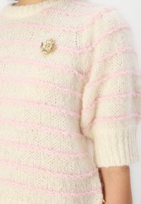 Weißer Strickpullover mit rosa horizontalen Streifen, kurzen Puffärmeln und einem goldenen Blumen-Anstecker als Akzent auf der Brust. Weiches, strukturiertes Material.