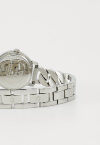 Reloj de acero inoxidable plateado con caja redonda, acabado suave y pulsera de eslabones. Presenta acentos de logo grabados en la correa.