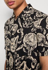 Schwarzhemd mit kurzen Ärmeln, dekoriert mit beigen Grafiken von Artischocken und Blättern, Knopfverschluss und klassischem Kragen. Weiche Textilstruktur.