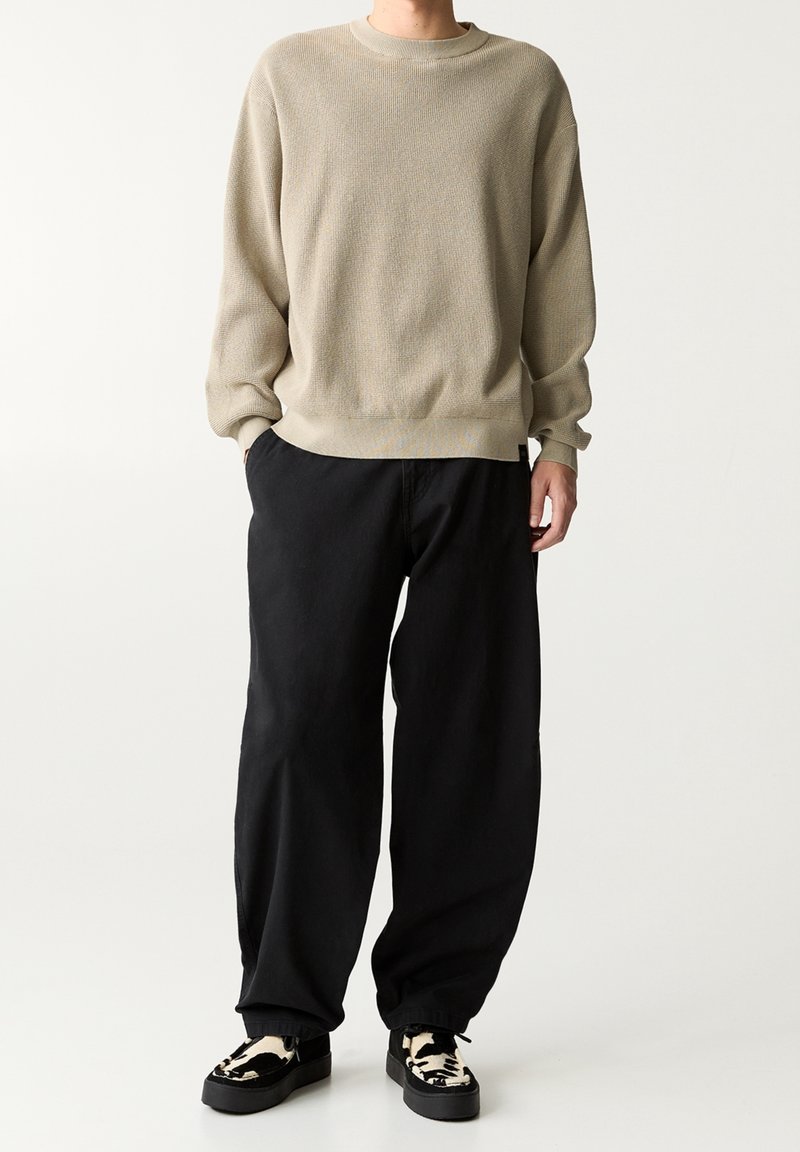 Personne portant un pull en maille beige, un pantalon noir ample et des chaussures slip-on à motif noir et blanc, debout devant un fond uni.