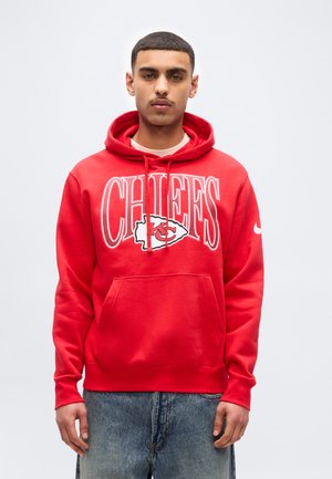 Mlad moški v rdeči kapucarji Kansas City Chiefs in modrih kavbojkah, stoji ob enostavni svetli ozadju.