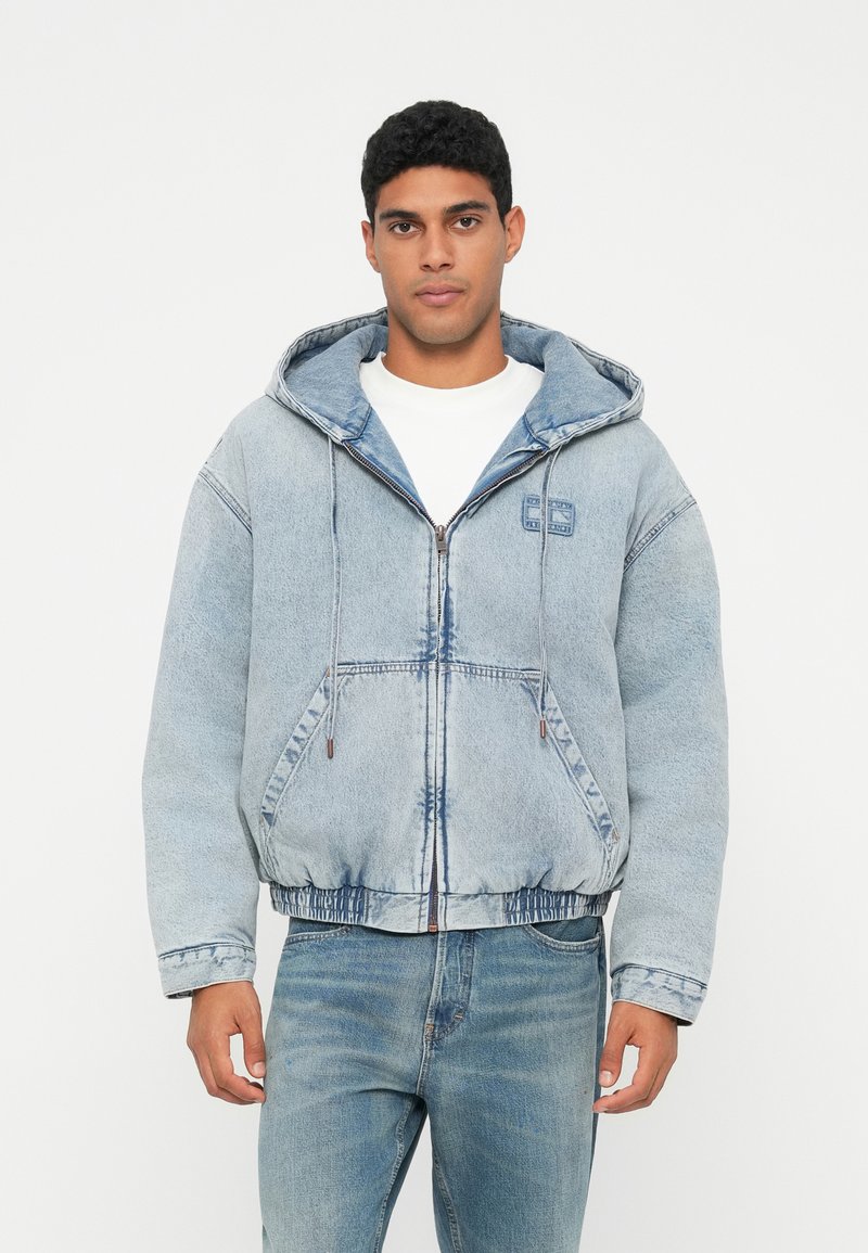Hombre vistiendo una chaqueta con capucha de mezclilla azul clara con cremallera sobre una camisa blanca y jeans azules, de pie frente a un fondo blanco liso.