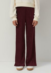Weite Hosen in Kastanienrot mit einer strukturierten Stoffoberfläche und vertikalen Rippungen. Kombiniert mit weißen Sneakers und einem cremfarbenen Pullover.