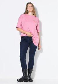 Camiseta de algodón rosa de manga corta con texto bordado en el pecho, combinada con jeans skinny oscuros y botas negras hasta el tobillo.