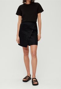 T-shirt noir associé à une jupe portefeuille texturée sombre arrivant aux genoux, avec des boutons sur le côté, et des sandales noires à lanières.