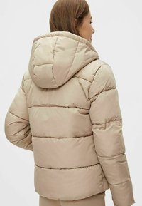 Beige polstret jakke med hette, med horisontal quilting og en lett cropped design. Glatt tekstur og lett materiale.
