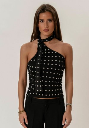 Mujer joven con cabello largo y castaño, vestida con una blusa negra de lunares de un solo hombro con un pañuelo anudado al cuello y pantalones negros, de pie en posición neutral.