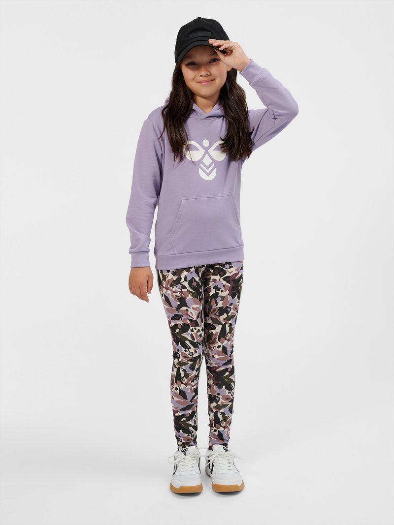Fille portant une casquette noire, un sweat à capuche lavande avec un logo blanc, des leggings camouflage aux tons violets et noirs, et des baskets blanches, debout sur un fond blanc uni.