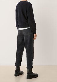 Osoba ubrana w czarne podwinięte dżinsy, czarny sweter, biały podkoszulek i czarne skórzane buty, stojąca na tle jednolitego jasnego tła.