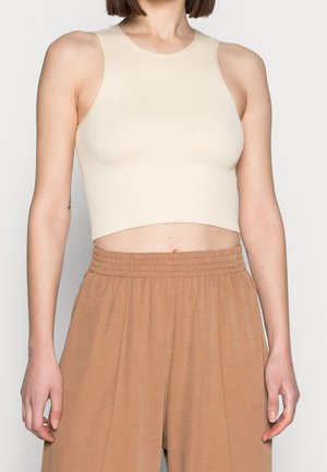 Kobieta w beżowym, bezrękawnym prążkowanym topie i luźnych brązowych spodniach z wysokim stanem, stojąca ze swobodnie opuszczonymi ramionami.