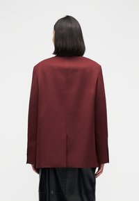 Bordeauxrood colbert met een rechte snit, knoopsluiting met twee knopen en een gestructureerde stof; heeft een enkele achtersplit en een gladde voering.