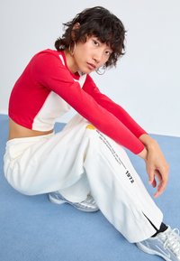 Červený dlouhý rukáv crop top spárovaný s bílými tepláky. Kalhoty mají potisk loga a černé detaily po straně. Na nohou má stříbrné tenisky.