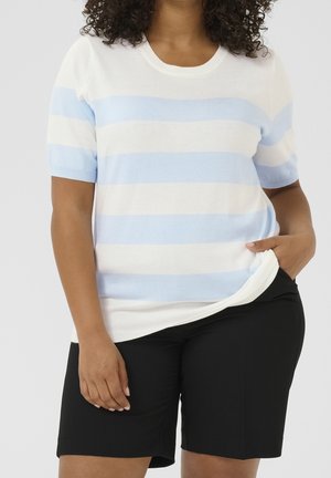 Person iført en hvid kortærmet sweater med lyseblå vandrette striber og sorte knæshorts, med den ene hånd i lommen.
