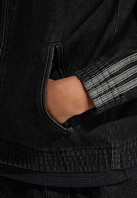 Svart jeansjacka med en dragkedjeförsedd ficka och tre texturerade grå ränder längs ärmslutet. Tyget verkar robust och lätt glansigt.