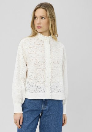 Blouse blanche avec broderie en dentelle florale complexe, col rond, boutonnage devant, et manches longues, associée à un jean en denim bleu.