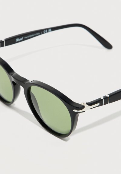 Γυαλιά ηλίου με μαύρο σκελετό, στρογγυλεμένους πράσινους φακούς και ασημένιες διακοσμητικές μεντεσέδες στους βραχίονες, τοποθετημένα σε λευκό φόντο.