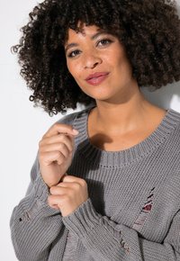 Grau gestrickter Pullover mit geripptem Halsausschnitt und Bündchen, mit abgewetzten Details und strukturiertem Stoff, gehalten von einer Person mit lockigem Haar.