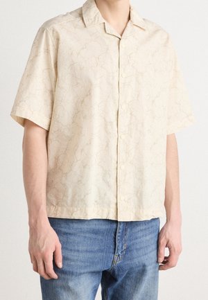 Man draagt een beige licht overhemd met korte mouwen en subtiel gemarmerd patroon, gecombineerd met een blauwe spijkerbroek, voor een effen achtergrond.