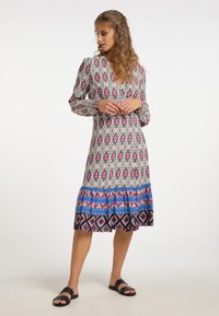 Femeie cu păr lung și ondulat, purtând o rochie midi cu model și mâneci lungi și sandale negre, stând în fața unui fundal alb simplu.