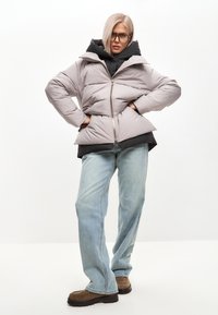 Lys grå pufferjakke med høj krave, lynlås og quiltet design, båret over en sort hættetrøje. Løse blå jeans og brune sko.