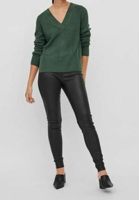 Zelený pletený svetr s výstřihem do V a žebrovanou strukturou, kombinovaný s černými legínami z umělé kůže. Obuv má špičaté špičky a nízké podpatky.