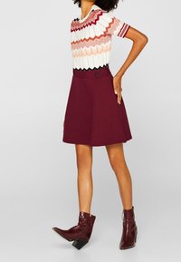 Korte mouw top met een rood, wit en roze zigzagpatroon; gecombineerd met een kastanjebruine flared rok met een zijknop. Bruine enkellaarsjes.