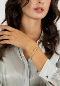 Bracciale in oro con due dettagli in cristallo, poggiato su una blusa argento texturizzata. Il bracciale presenta un design elegante e minimalista.