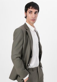 Olive green suit with a slim fit, featuring a single-breasted jacket, notched lapels, and matching trousers. White shirt underneath.