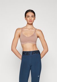 Lys brun sport-bh med tynne stropper, glatt tekstur, minimalistisk design og en liten logodetalj. Kombinert med høyt livet blå tights.