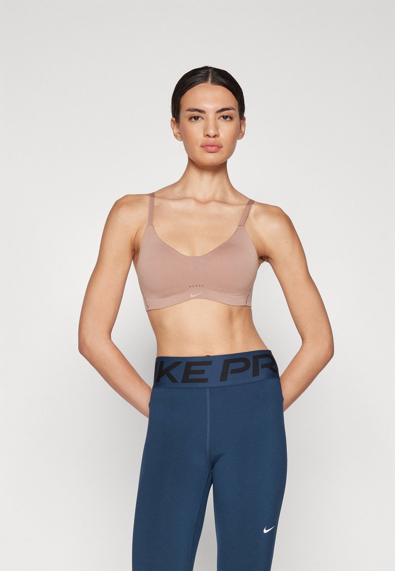 Lys brun sport-bh med tynne stropper, glatt tekstur, minimalistisk design og en liten logodetalj. Kombinert med høyt livet blå tights.