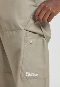 Beige Hosen aus leichtem Stoff mit einer seitlichen Tasche mit Knopfverschluss und einem weißen Logo am unteren Bein.