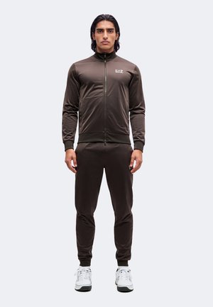 Homme debout portant un survêtement zippé marron foncé avec le logo EA7 et des baskets blanches sur un fond clair uni.