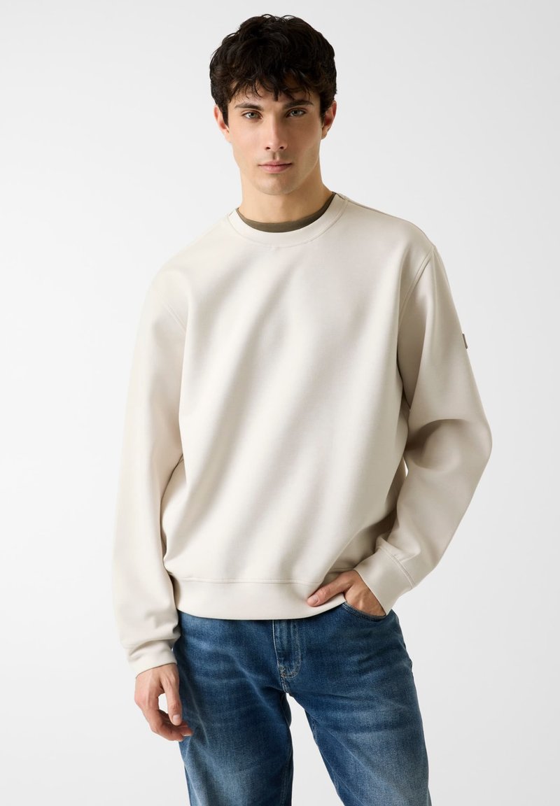 Sweat-shirt beige à manches longues, col rond et poignets côtelés. Porté avec un jean bleu. Design simple, texture lisse, sans motifs visibles.