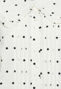 Vit texturerad skjorta med svarta prickar, med en rynkad krage och små pärlknappar längs framsidan.