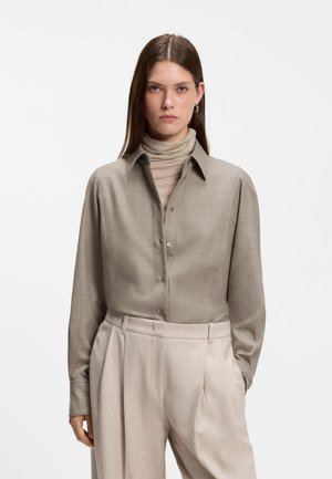 Kvinde med glat brunt hår iført beige rullekrave under en taupe knapbetjent skjorte med lysebeige plisserede bukser, står med hænderne i lommerne.