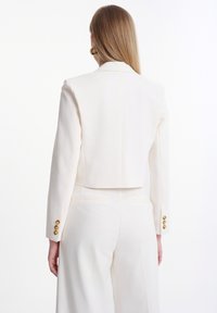 Blazer bianco corto con maniche lunghe, caratterizzato da tre bottoni dorati su ciascun polsino, tessuto liscio e design sartoriale.