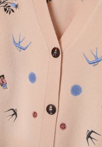 Roosa villane kardigan tikanditega, mis kujutavad linde, päikesi ja kujusid; omab V-kaelust, soonikulisi servi ja ümmarguseid nööpe.