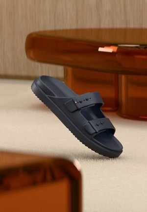 Vienas juodas šlepetės tipo sandalėlis su dviem reguliuojamomis dirželiais, gulintis ant smėlio spalvos kilimo šalia rudų permatomų stalo kojų.