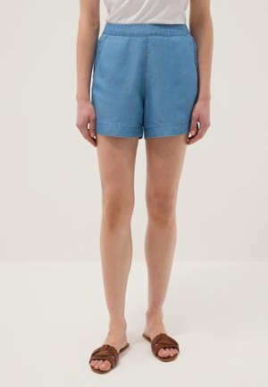 Frau trägt hellblaue Denim-Shorts, weißes T-Shirt und braune geflochtene Sandalen vor einem einfarbigen hellen Hintergrund.