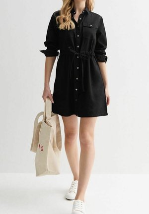 Žena v černých košilových šatech s ohrnutými rukávy, držící béžovou tašku a chodící v bílých teniskách na jednobarevném pozadí.