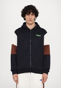 Hooded zip-up sweatshirt in navy with brown and white sleeves. Features a green logo on the chest and sturdy cuffs. Soft fabric texture.