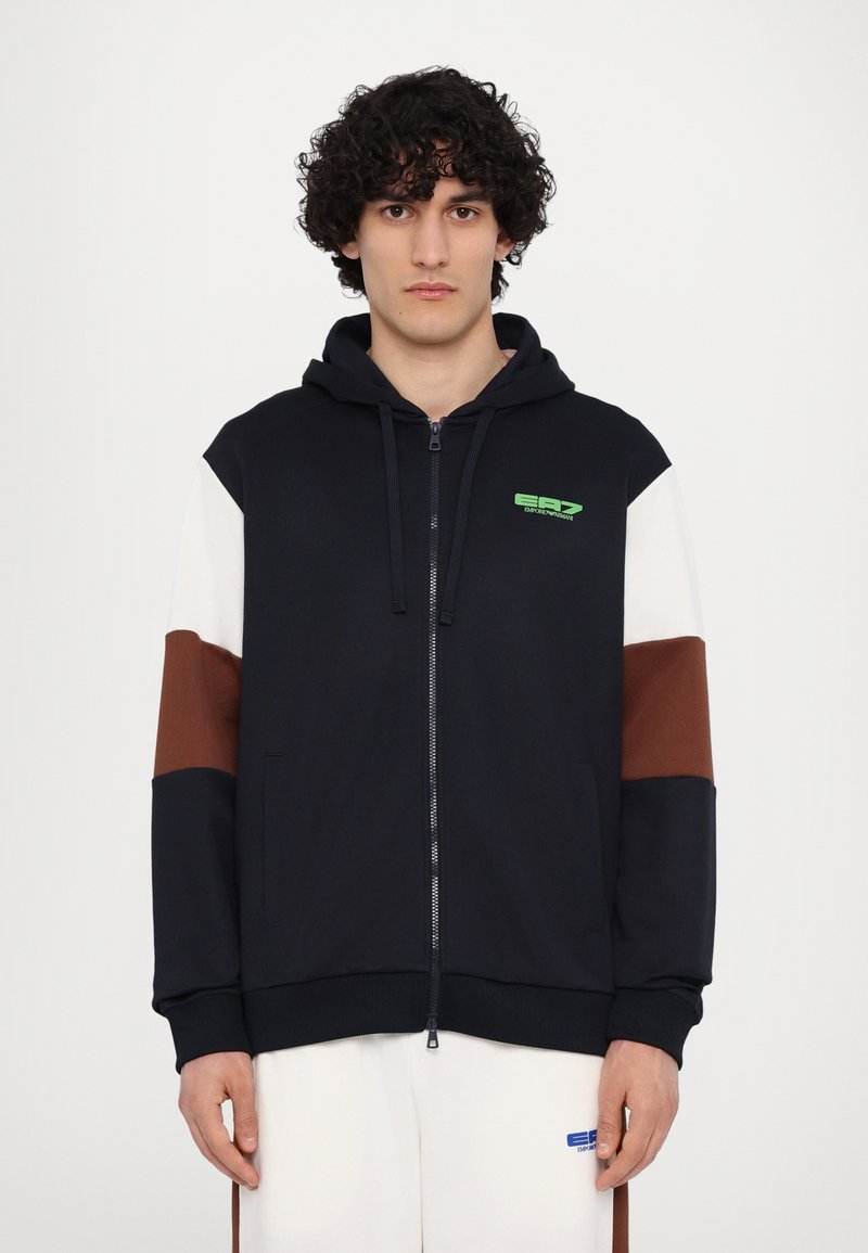 Hooded zip-up sweatshirt in navy with brown and white sleeves. Features a green logo on the chest and sturdy cuffs. Soft fabric texture.