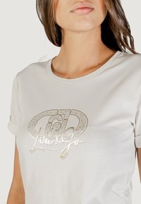 Donna con una t-shirt bianca con maniche arrotolate, decorata con una grafica metallizzata oro e la scritta "Liu Jo Milano" sul petto.