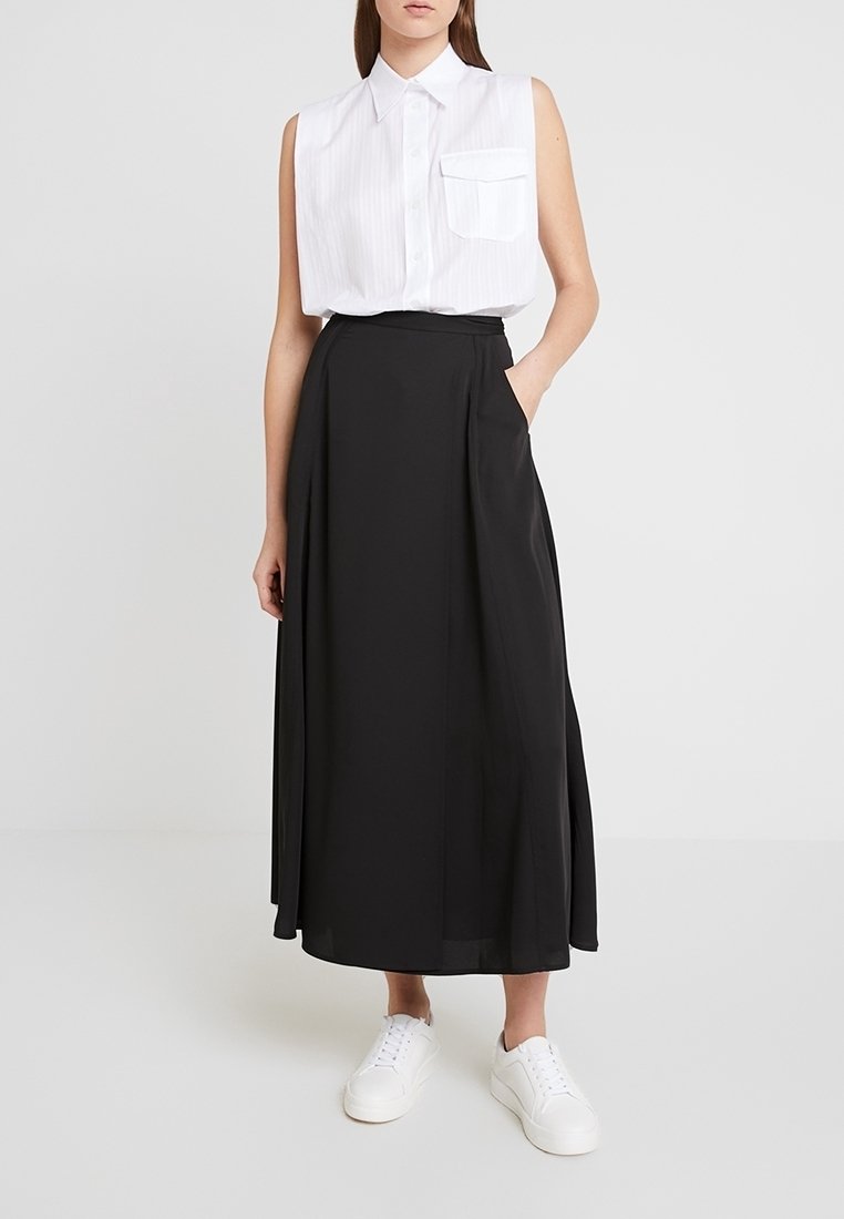 Donna che indossa una camicia bianca senza maniche, una lunga gonna nera a vita alta con tasche e sneakers bianche su sfondo bianco.