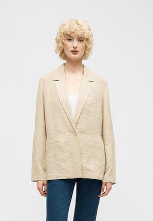 Blonde Frau mit lockigem Haar trägt ein beiges, strukturiertes Blazer über einem weißen Oberteil und blauen Jeans und steht vor einem schlichten, hellen Hintergrund.